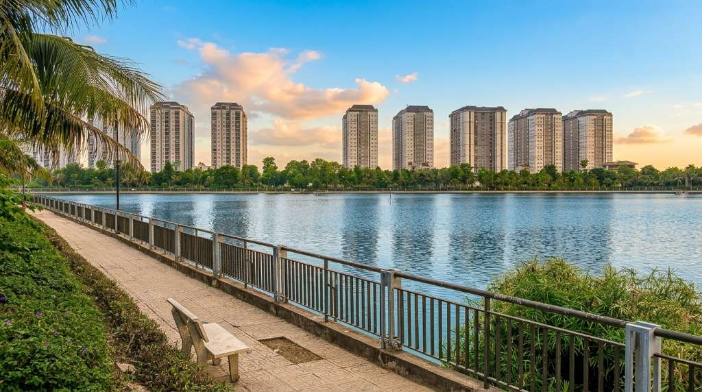 Ảnh thực tế KĐT Thanh Hà mùa hè 2025, con đường nội khu rợp bóng cây xanh đã trưởng thành hai bên, phía xa là hồ cảnh quan nước trong xanh phản chiếu tòa chung cư, có vài người dân đi dạo buổi chiều, ánh nắng xuyên qua tán lá tạo hiệu ứng đốm sáng trên mặt đường, góc chụp thẳng dọc con đường tạo chiều sâu, tông màu xanh mát