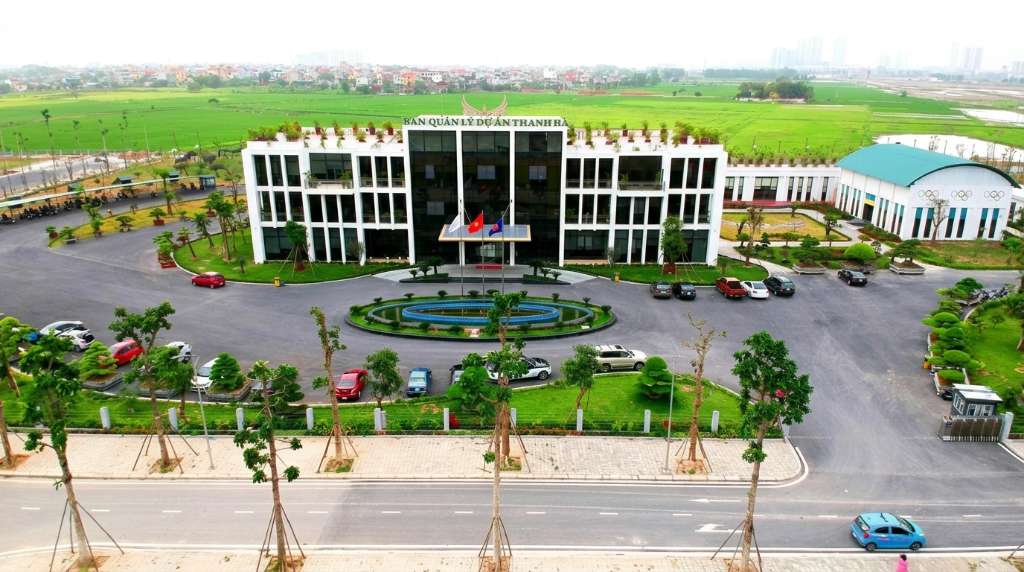 Tay một người cầm checklist in trên giấy A4 với các mục đánh dấu tick bằng bút đỏ (kiểm tra PCCC, sổ hồng, tầng hướng, chất lượng xây dựng), phía sau mờ nhòe là sảnh tầng 1 chung cư Thanh Hà với cửa kính lớn nhìn ra khuôn viên cây xanh, ánh sáng tự nhiên chiếu vào, góc chụp close-up tay và tờ giấy, bokeh nền đẹp