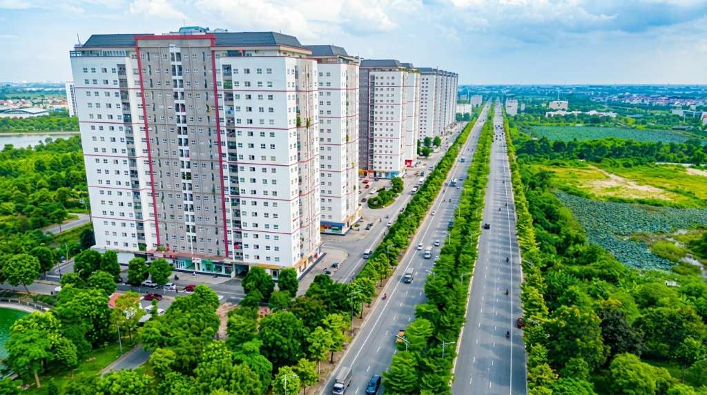 Khu vực sảnh chính tòa chung cư HH03 Thanh Hà vào buổi sáng, cư dân gồm các gia đình trẻ bế con nhỏ và nhân viên văn phòng đang chờ thang máy, ánh sáng tự nhiên chiếu qua cửa kính lớn, bảng thông báo cư dân treo trên tường, góc chụp ngang tầm mắt, phong cách ảnh báo chí đời thường